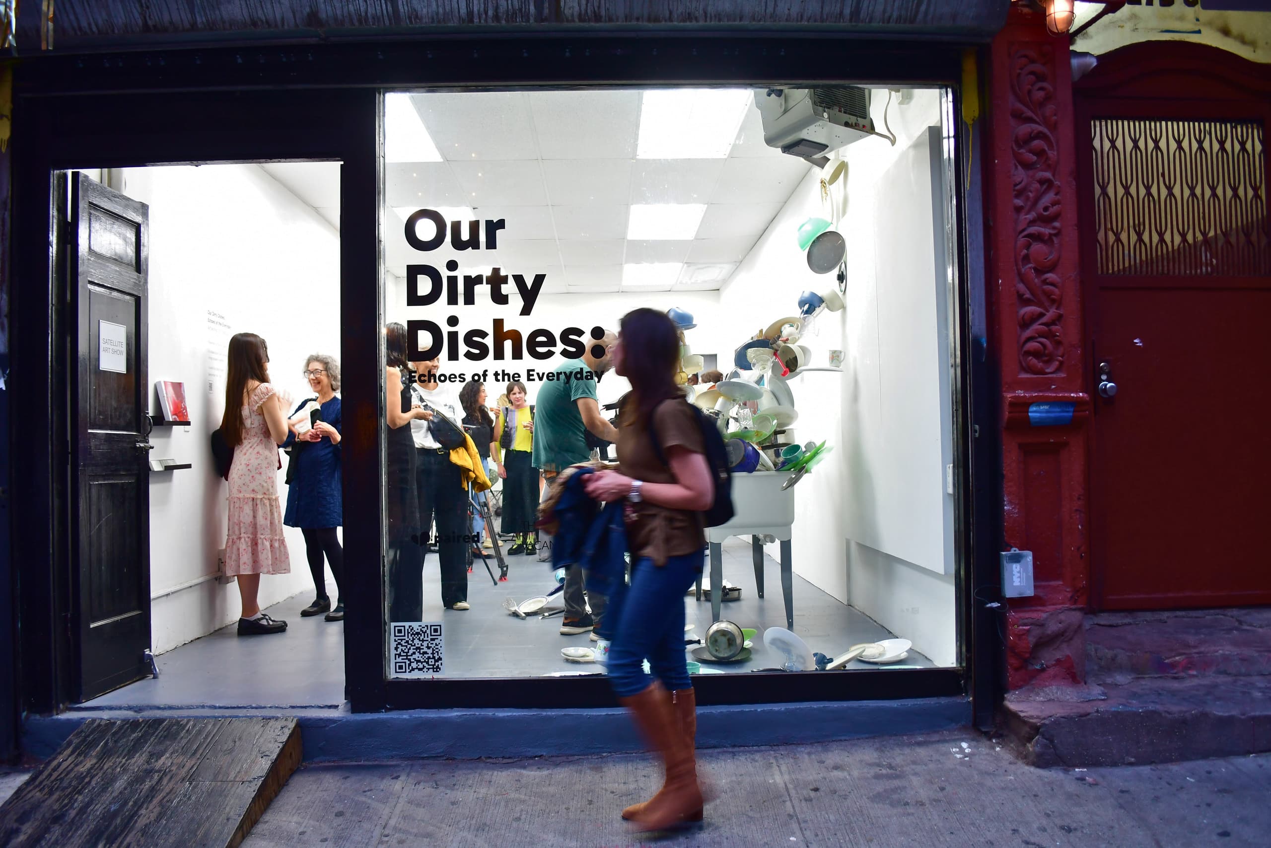 Our Dirty Dishes - NYC exhibition from Paired. The image shows a crowd of people inside an art gallery, with 'Our Dirty Dishes - Echoes of the Everyday' on the window. Behind that is a tall structure of dishes. A person walks past outside the window.